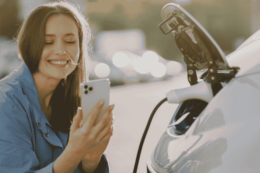 Mulher sorridente usando celular enquanto carro elétrico carrega, simbolizando praticidade na escolha do modelo mais barato em 2025.