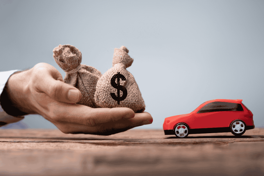 Mão segurando sacos de dinheiro ao lado de carro em miniatura vermelho, simbolizando custo total de propriedade e economia na compra de carros bons e baratos.