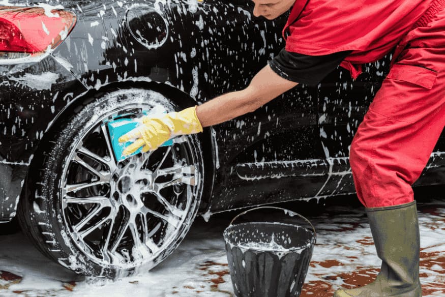 Pessoa vestindo macacão vermelho e luvas amarelas lavando a roda de um carro preto com uma esponja azul, rodeada de espuma, com balde ao lado.