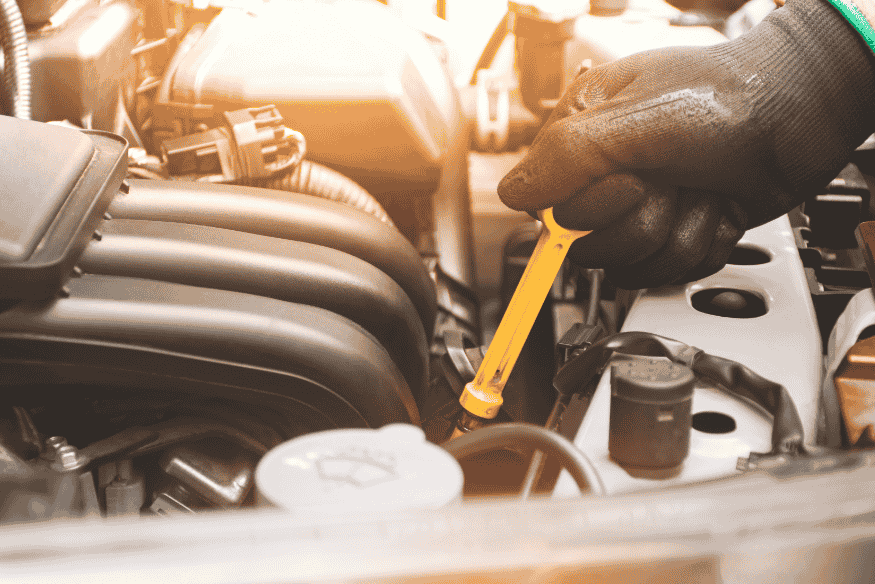 Mão com luva segurando uma vareta de óleo de cor amarela dentro do compartimento do motor de um carro, com luz suave iluminando a cena.