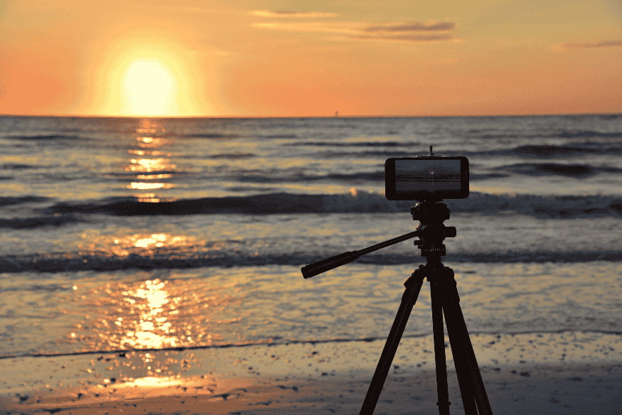 Celular em tripé fotografando o pôr do sol na praia, mostrando a importância da estabilidade para fotos em RAW