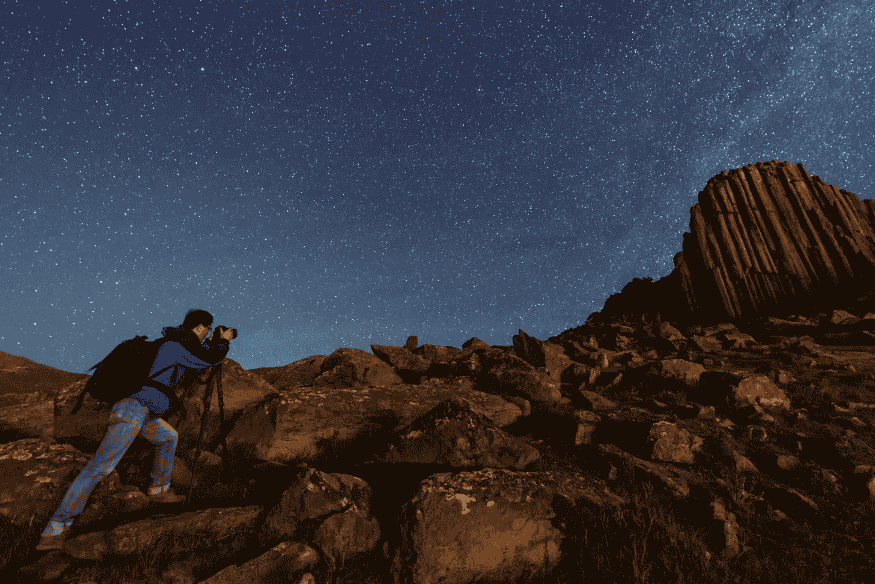 Fotógrafo usando câmera em paisagem rochosa sob céu estrelado, mostrando a importância da escolha da lente na astrofotografia conforme a Escala de Bortle
