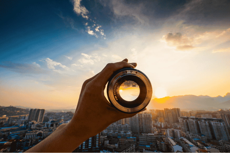 Pessoa segurando lente de câmera diante do horizonte urbano ao pôr do sol, ilustrando o uso de lente grande angular para fotografar o céu
