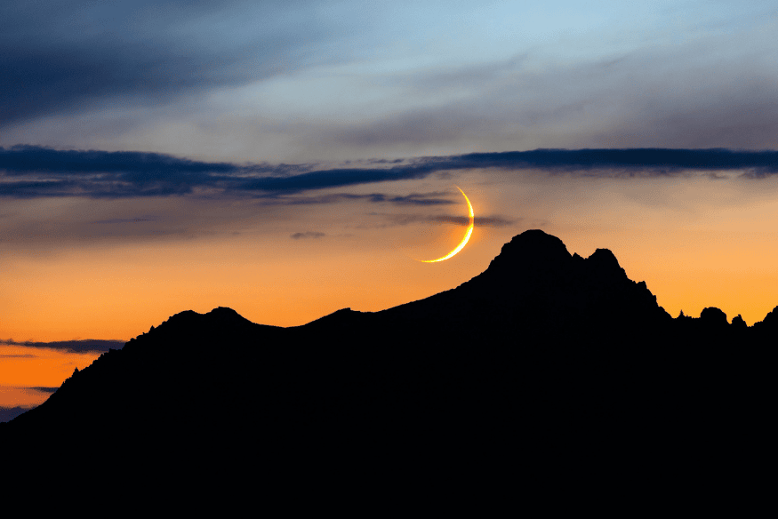 Lua crescente alaranjada próxima ao horizonte e atrás de montanhas ao entardecer, ilustrando como a baixa altitude no céu intensifica a mudança de cor da Lua