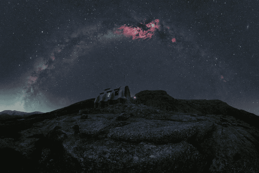 Arco da Via Láctea sobre observatório em paisagem escura, destacando a mudança de posição da galáxia ao longo do ano