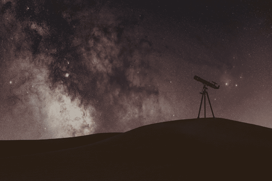 Telescópio em silhueta sob o arco brilhante da Via Láctea no céu noturno, mostrando que o centro galáctico pode ser visto mesmo sem equipamento avançado