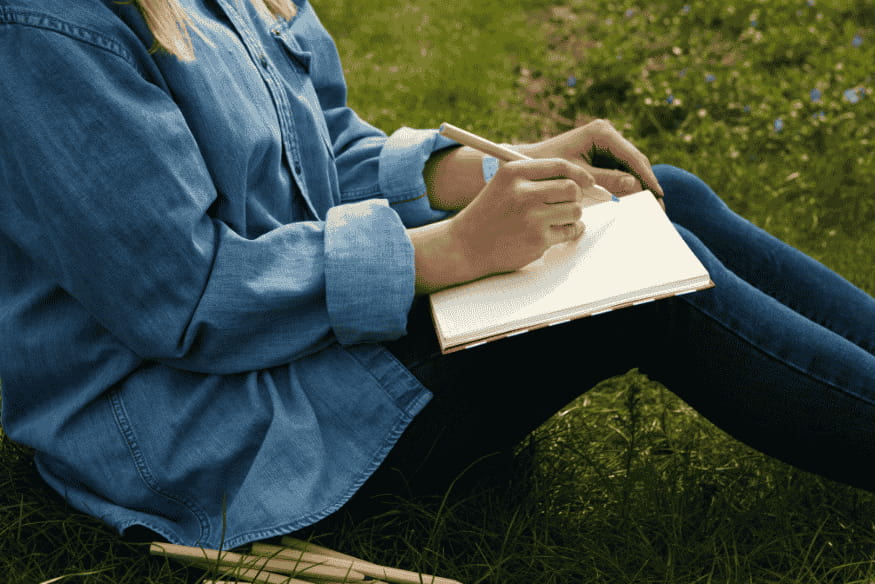 Pessoa escrevendo em um caderno simples no gramado, mostrando que um diário de observação astronômica pode começar com anotações feitas à mão