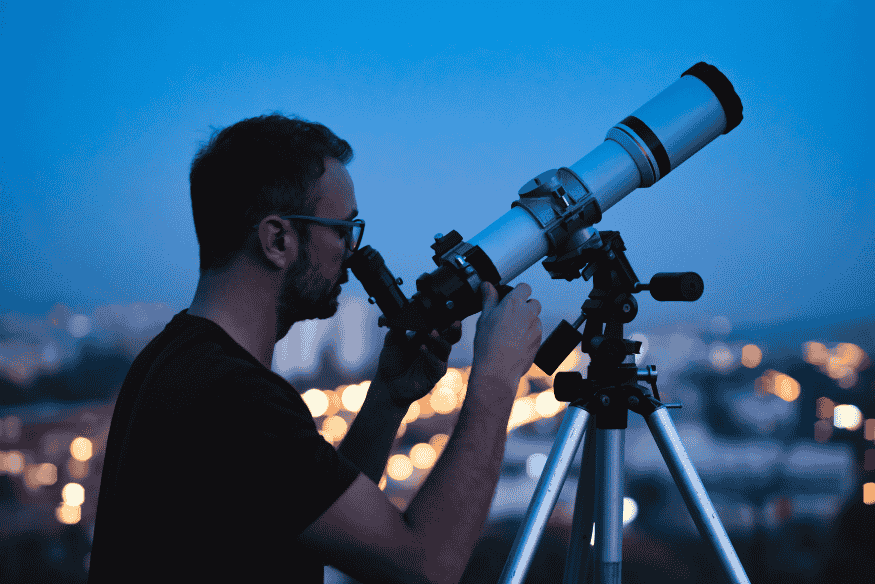 Observador usando telescópio ao anoitecer na cidade, momento ideal para registrar detalhes em um diário de observação astronômica