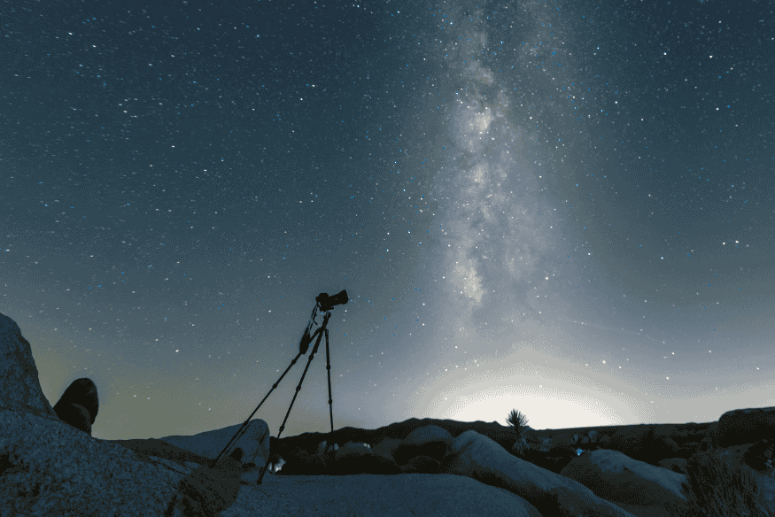 Tripé com câmera apontada para a Via Láctea sobre paisagem rochosa, ilustrando como o pós-processamento na astrofotografia pode valorizar detalhes do céu e do primeiro plano